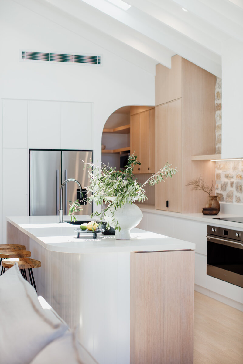 Modern kitchen with white countertops, wooden cabinets, and stainless steel appliances, crafted by a full-service interior design Newcastle expert. A vase of greenery adds a touch of nature to the sleek island centerpiece.