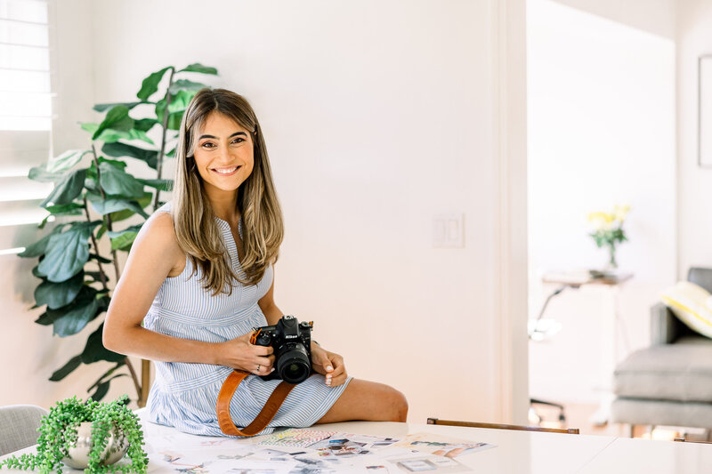 Greenwich, Connecticut family photographer smiling in her bright, cozy studio space, holding her camera and surrounded by printed memories. She captures playful, love-soaked moments for families who dance in the kitchen, cuddle close, and want to remember it all—just as it felt.