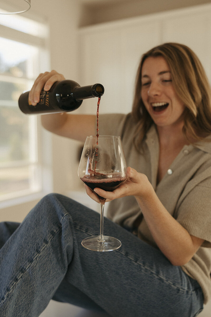 Wedding photographer pours red wine into her glass as she sits on her kitchen island