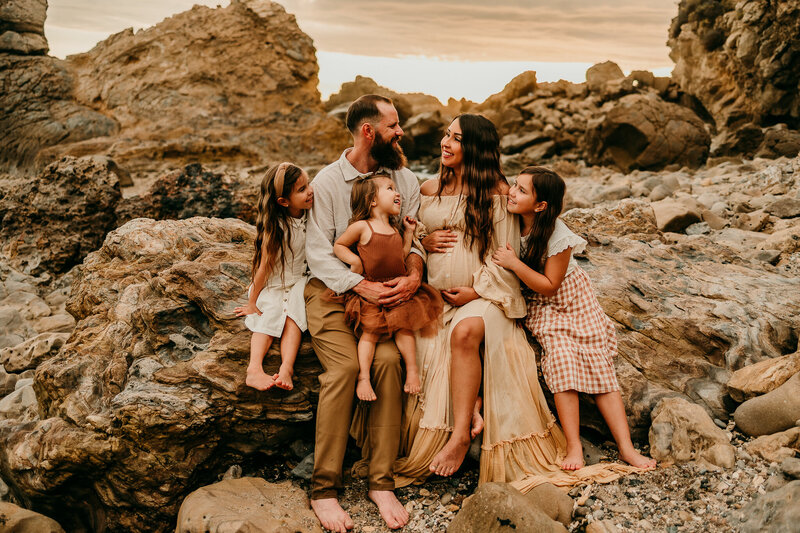 A growing family with four children cuddled together on coastal rocks during a golden-hour maternity and family session in Southern California, styled in boho neutrals