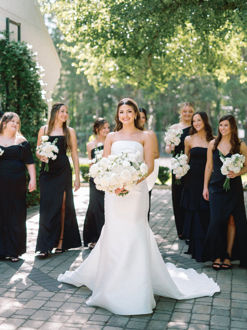 Modern bride walking with bridesmaids in black dresses with elegant and a bow on her dress with white bouquet