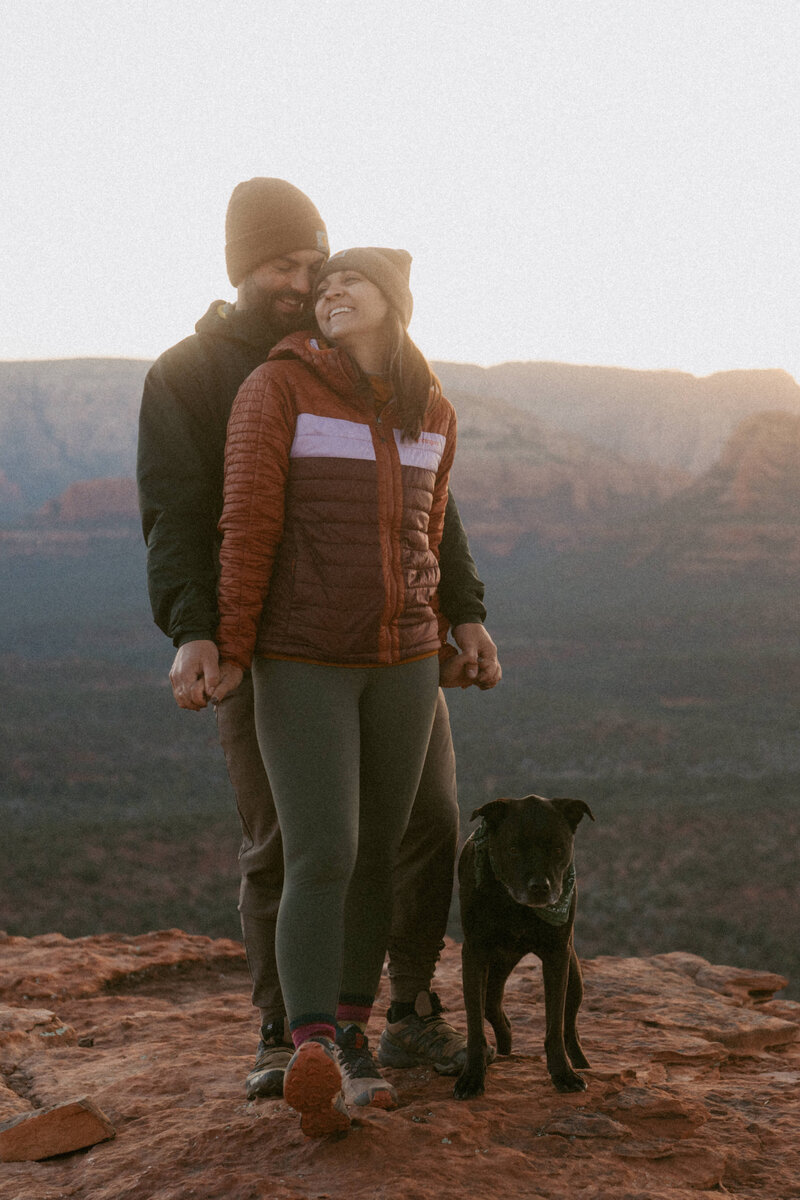 Smiling couple and their dog snuggling together at sunrise on the edge of a cliff in Sedona, Arizona.