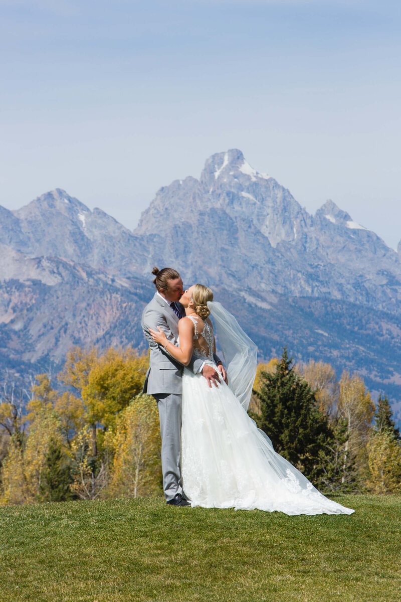 A newlywed couple in front of the stunning Grand Teton mountains at their wedding, with the bride’s veil gently flowing in the breeze and her flowing gown beautifully highlighted against the natural landscape.