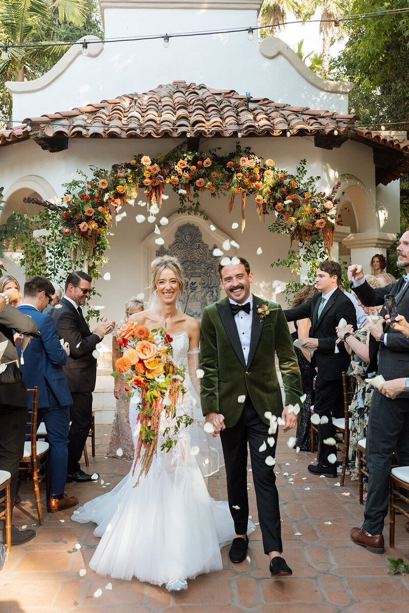 Young couple walking down wedding aisle at Rancho Las Lomas with petals being tossed and bright fall colored florals in a white lace dress and velvet green suit 