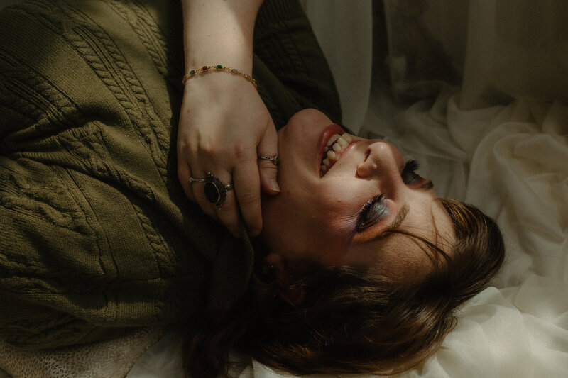 PA Wedding/Boudoir Photography. Photographer laying down with her eyes closed and laughing. She's wearing a green knit sweater and has colorful makeup on her eye lids.