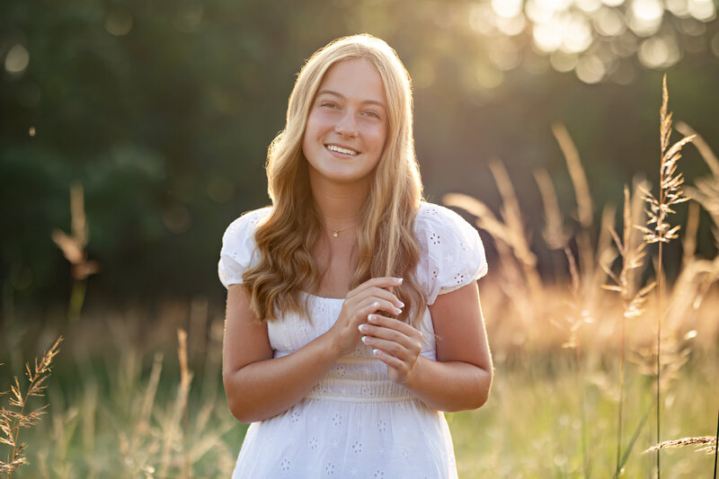 a young woman stands in a field of grass with golden sunshine behind her as she plays with a strand of hair in a white dress