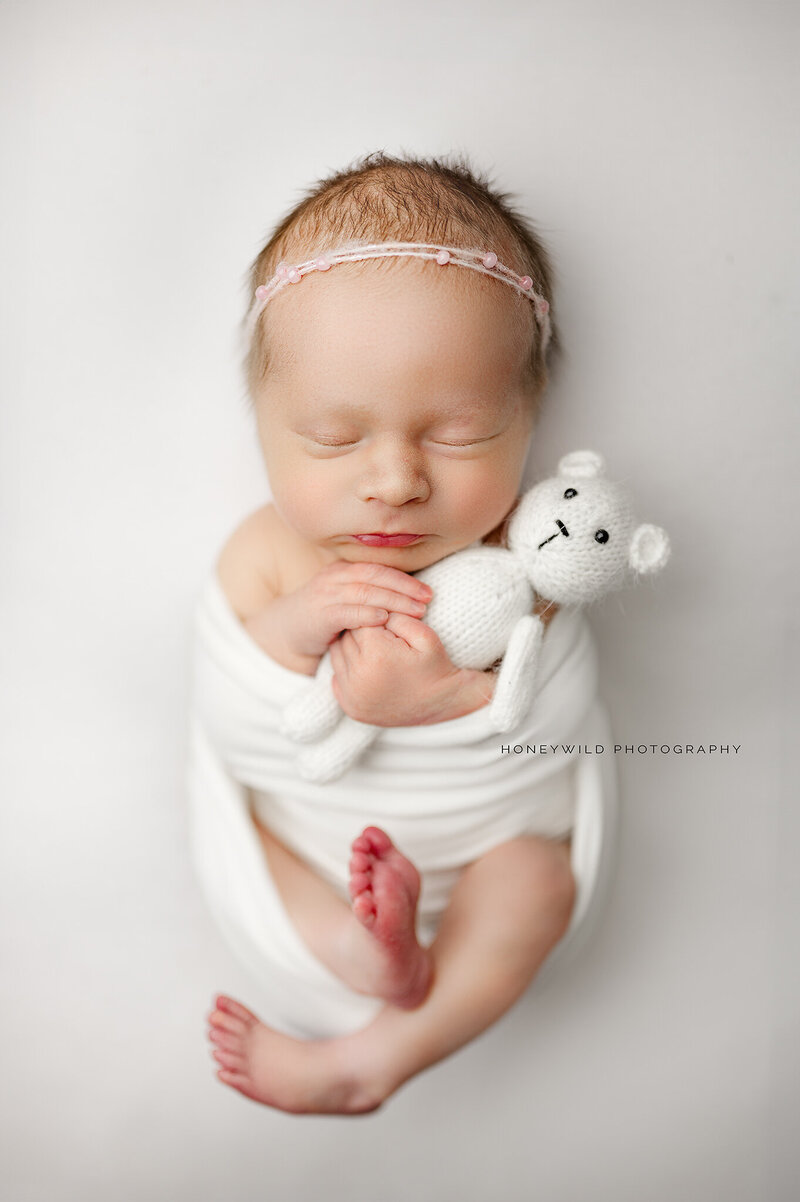 Newborn baby girl swaddled in white, holding a small knitted teddy bear, captured during a professional newborn photography session in Grand Rapids, Michigan