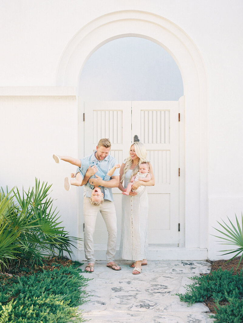 Mom and dad holding their two children and laughing in front of a white door and building