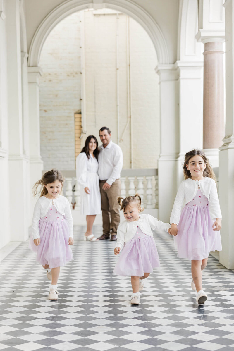 Family portraits of mum and dad in the background together watching their 3 children in purple dresses walk towards the camera together in the foreground.