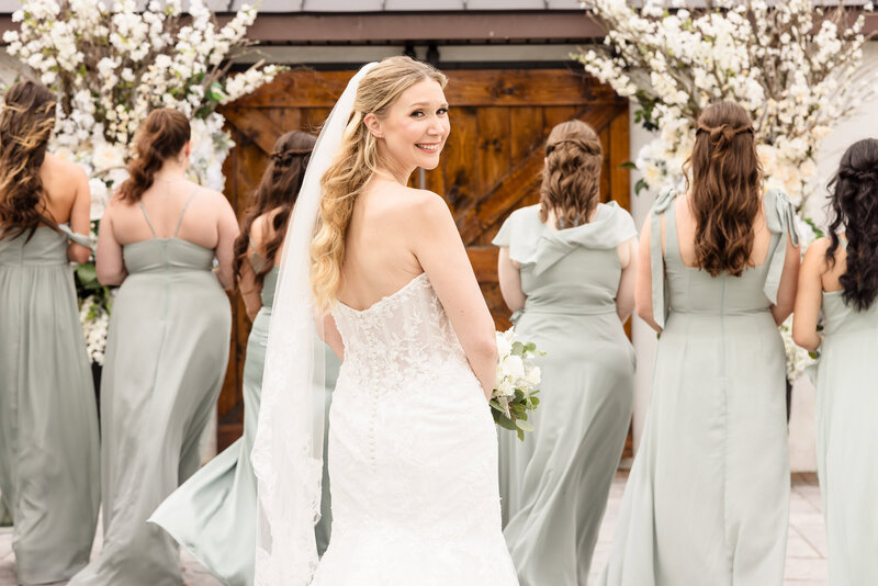 Smiling bride glancing over her shoulder in front of her bridesmaids in sage green dresses on wedding day.