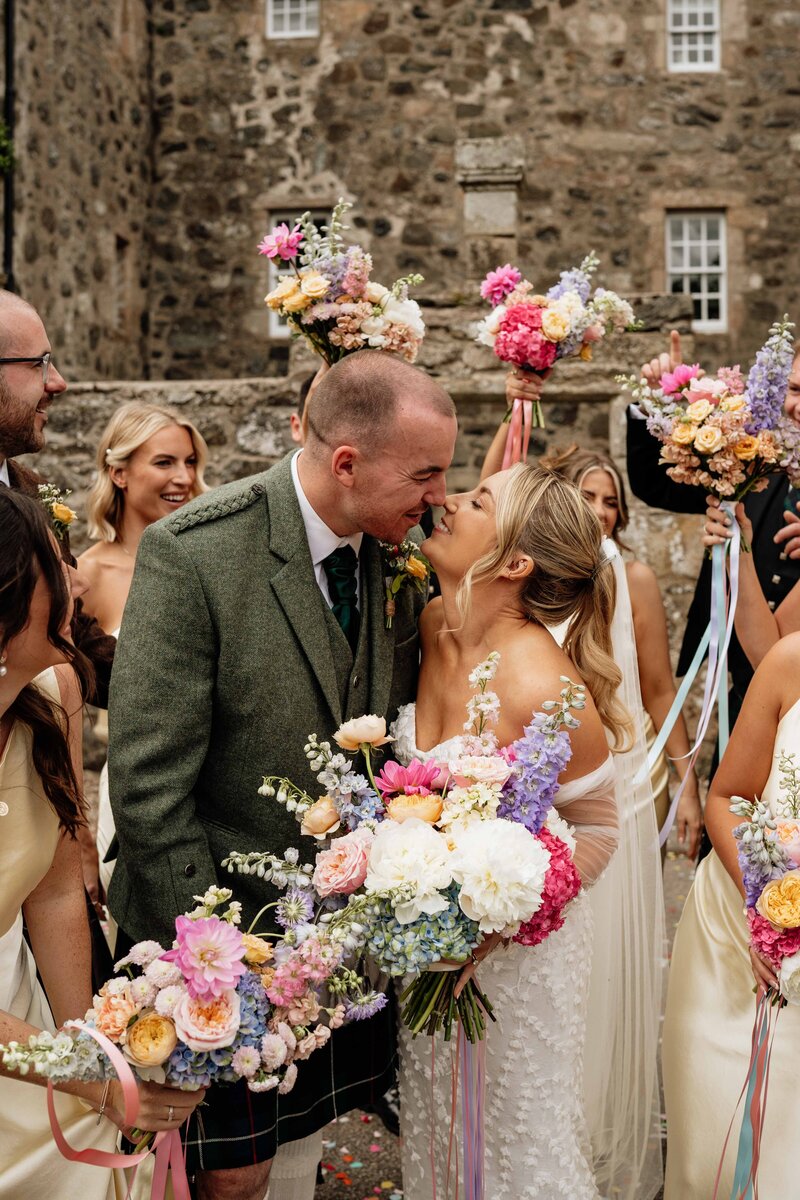 
A joyful photograph of a wedding party surrounding a newly married bride and groom after their wedding. Colourful bouquets and clothing make the photograph feel full of life as the couple kiss.