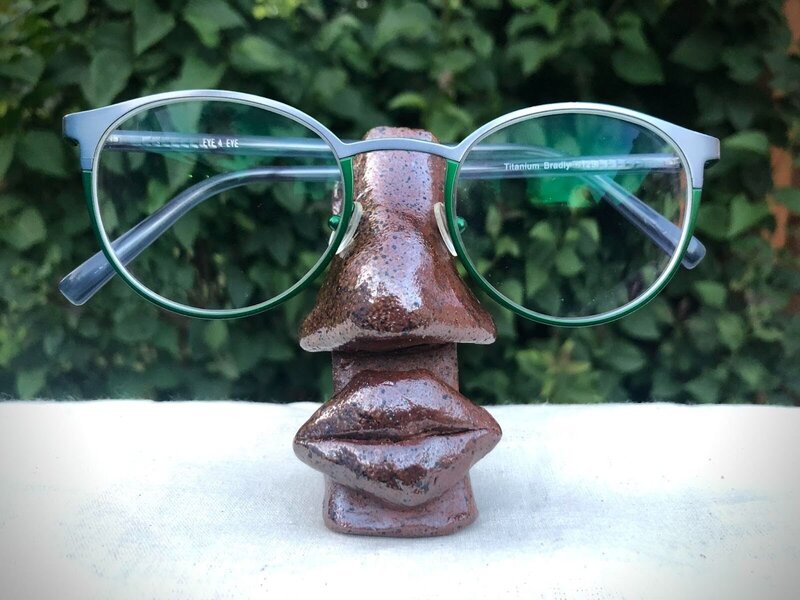Dark brown pottery shaped into a human nose and mouth, with blue and green rimmed glasses sitting atop the nose