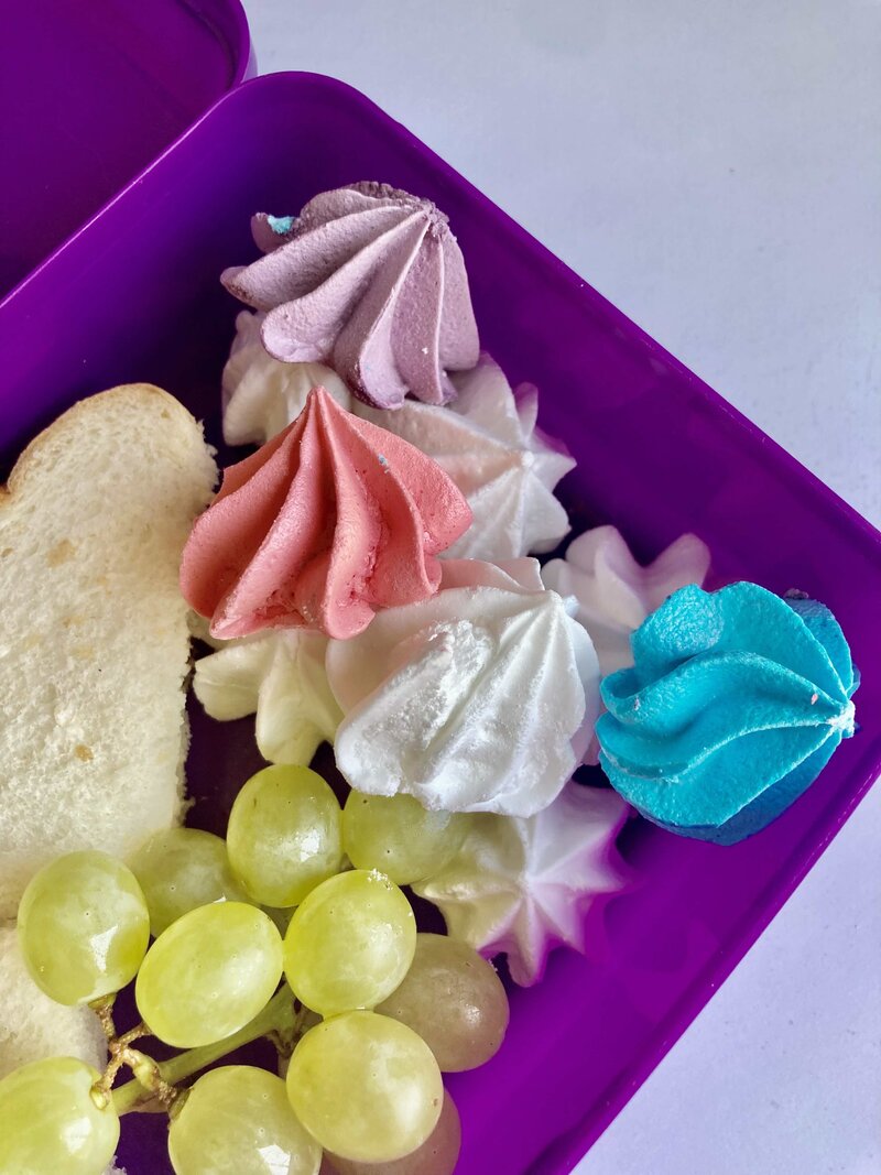Purple lunchbox with meringue pink, purple, blue and white meringue rosettes, green grapes, and a sandwich