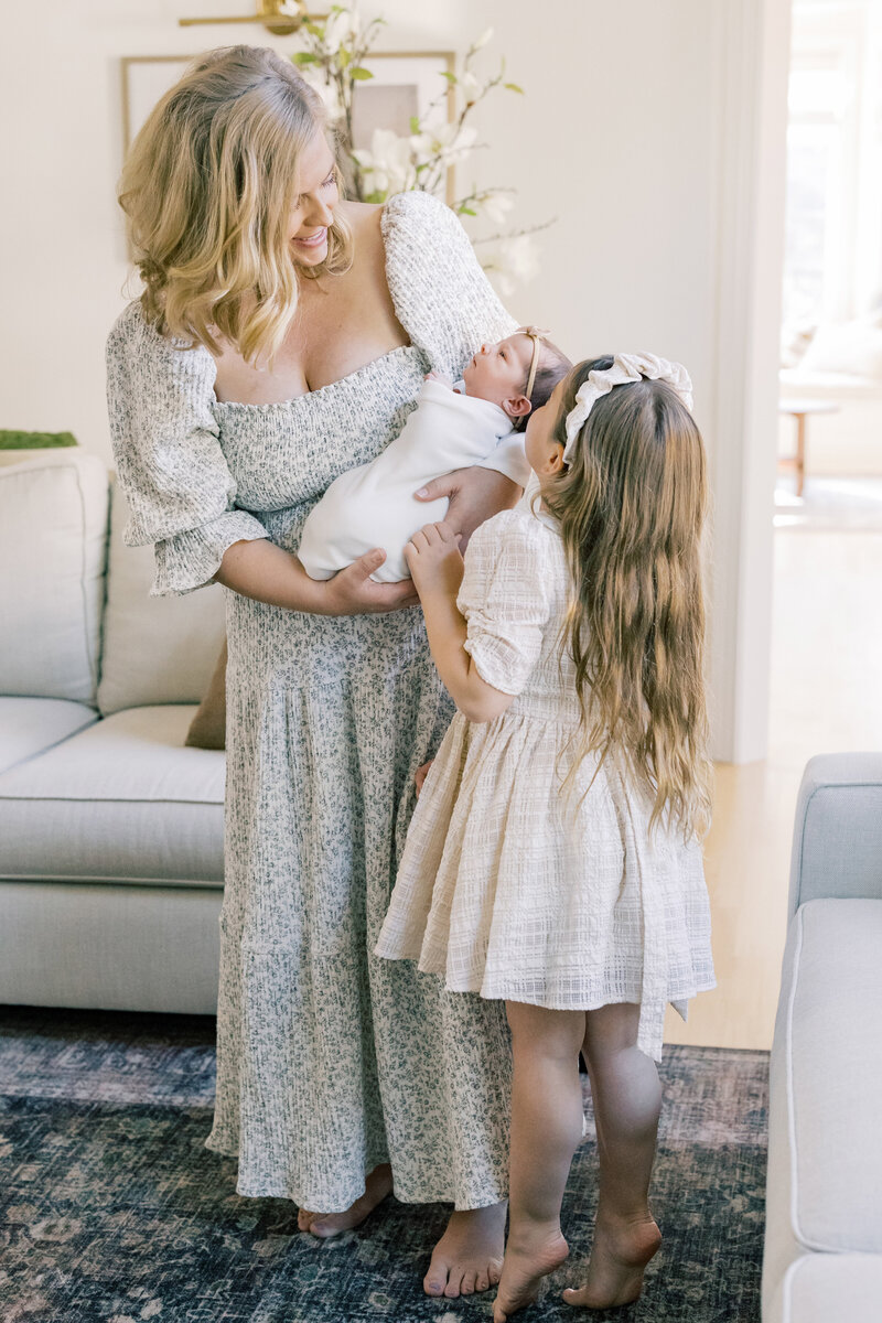 Mother holding newborn while big sister reaches in, captured in soft light during an in-home family session.