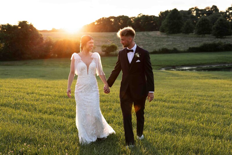 bride and groom walking across green grass while smiling at each other with the sun setting behind them