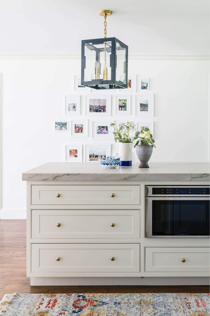Milton kitchen designer renovates a white kitchen with blue lantern pendants, marble counters, an under-counter microwave, and framed personal art on white walls.