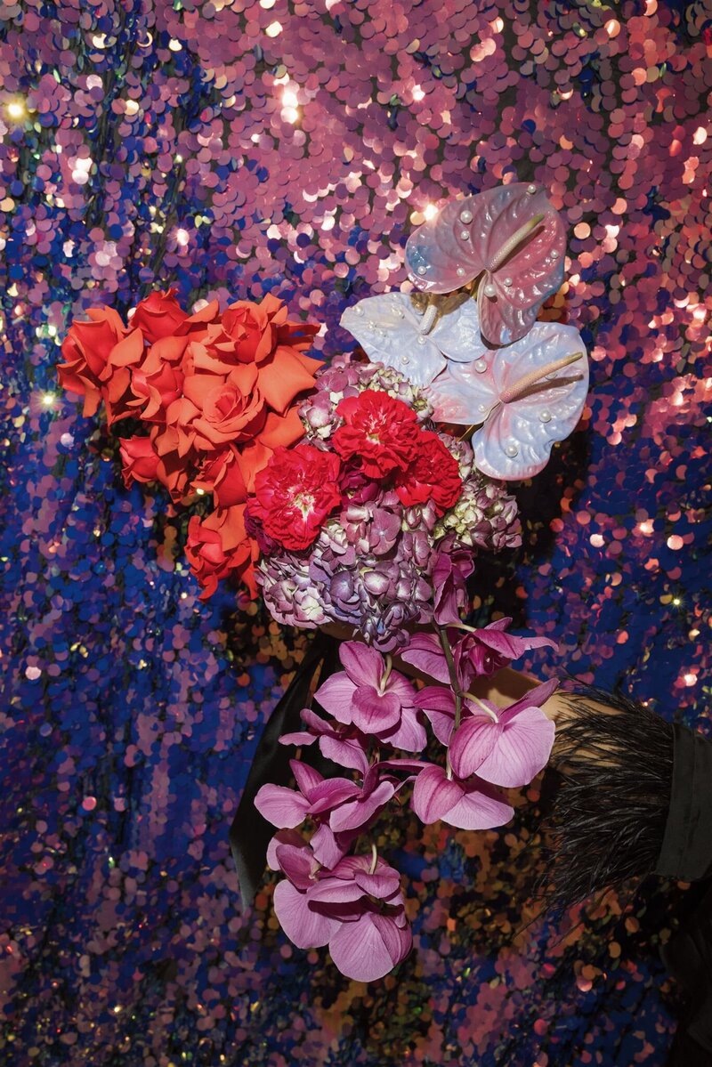 Elaborate colorful modern bridal bouquet with pearl iridescent anthuriums, red roses, red carnations, purple hydrangeas, and purple lilies in front of an iridescent sequins backdrop 