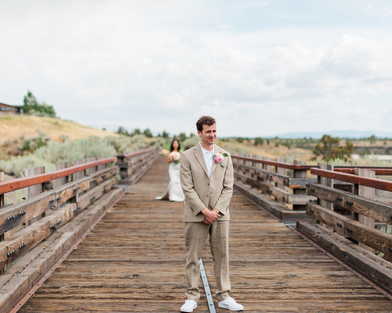 Groom in tan suit is standing in the middle of a wooden bridge, looking at the camera, waiting with his hands folded in front of him. His bride is in the background behind him, walking towards him for their first look before their wedding ceremony. He cannot see her yet.
