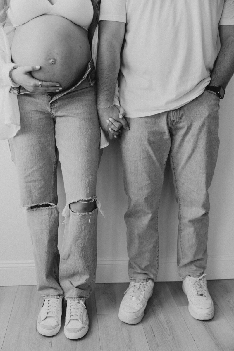 Black and white maternity photo of a couple in ripped jeans and sneakers, holding hands and embracing the baby bump during a cozy in-home session by a Michigan Ohio maternity photographer.