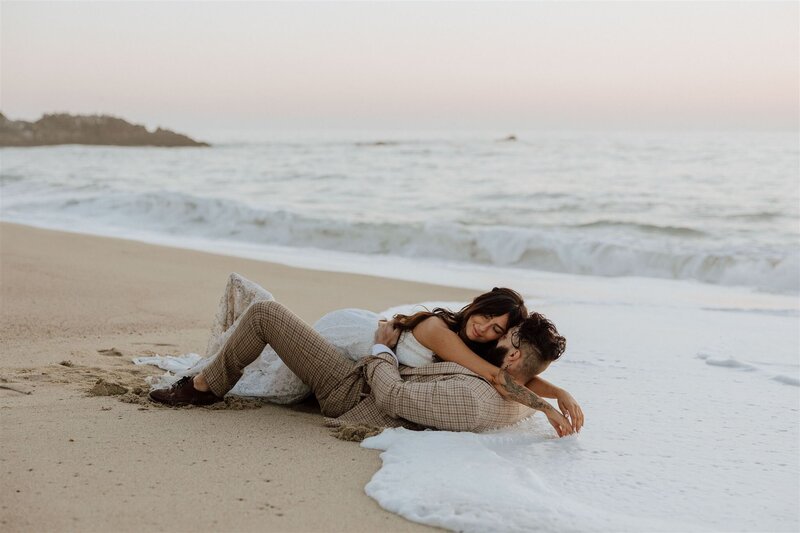 bride and groom laying in the sand on a beach with ocean waves gently surrounding them