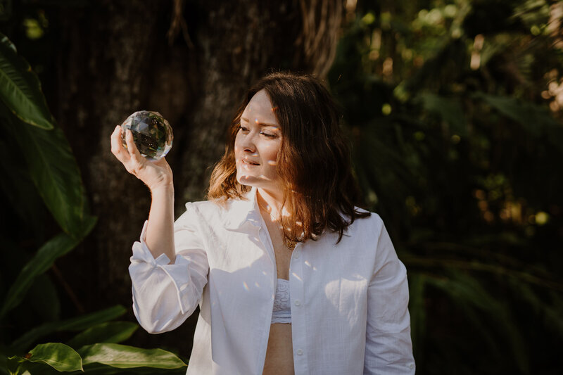 Brandi of the Intuitive Momma stands outdoors in a white button down looking into a crystal ball reflecting the sun