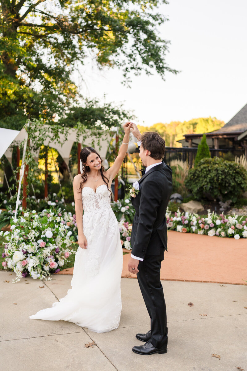 A groom holds his bride's hand and spins her in front of an elaborate pastel floral arrangement leading into their tented reception