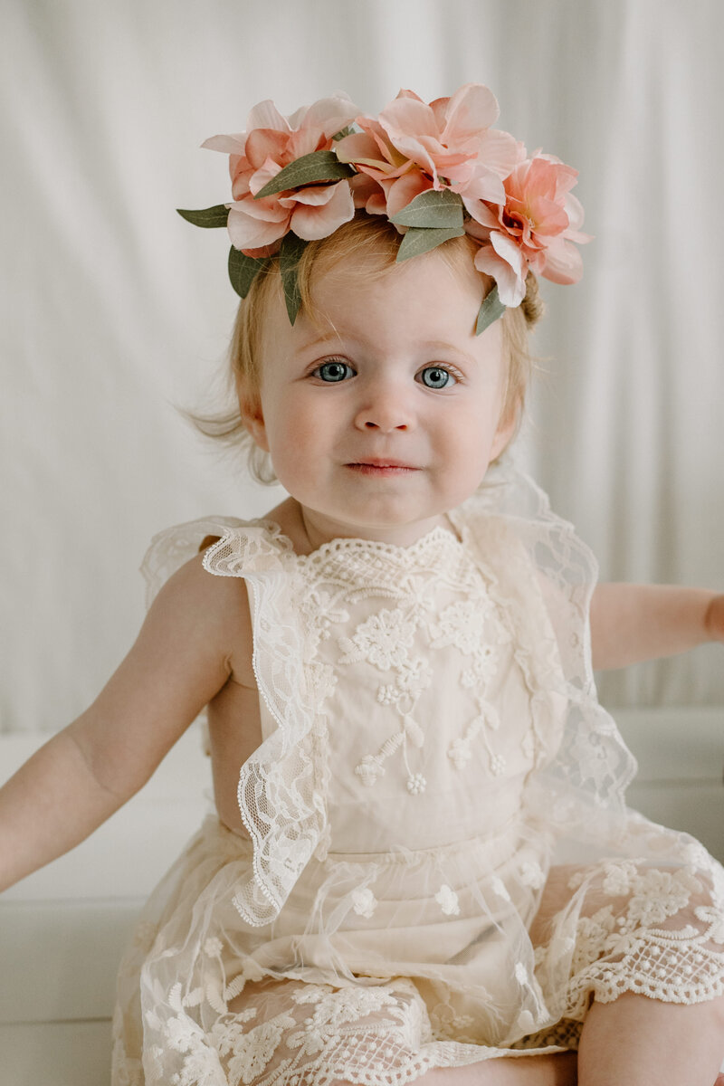 Baby girl in white lace with pink floral crown sitting in white studio in Severn Maryland photographed by Bethany Simms Photography
