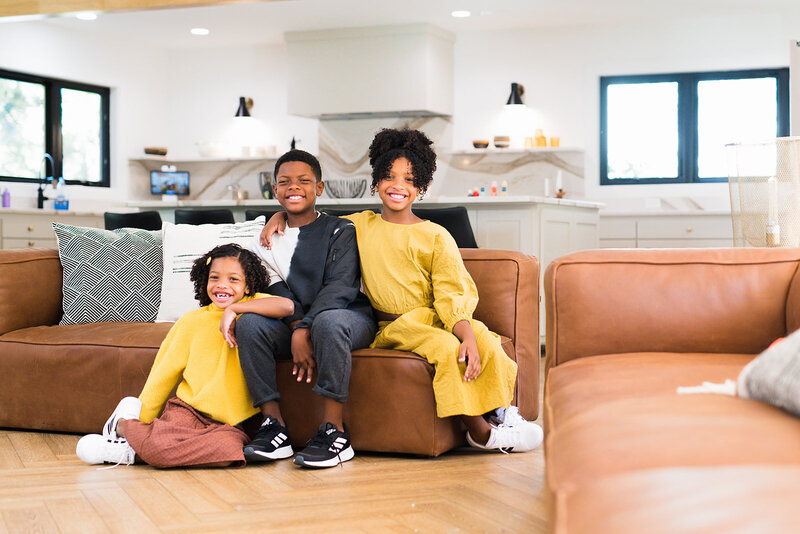 Three children smile at the camera from their living room for a family photoshoot. They sit on a leather couch.