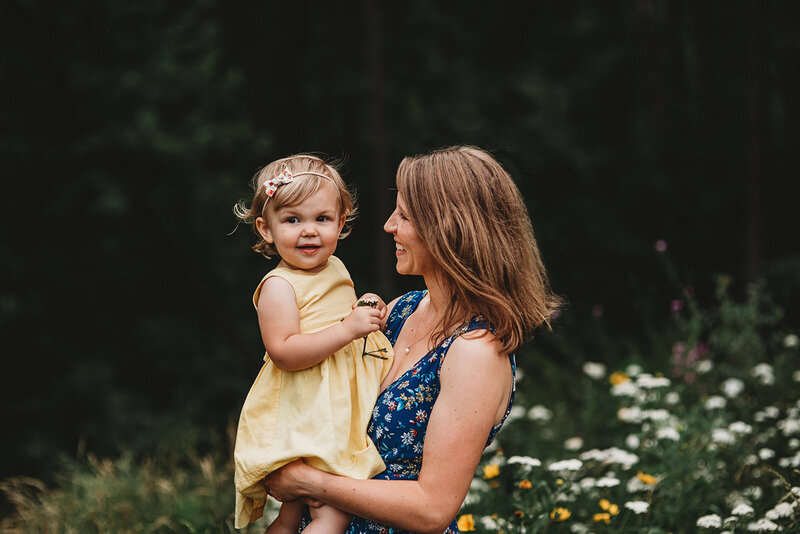 Baltimore photographers  photographs mother in a blue dress holding her daughter who is in a yellow dress and holding a flower with a field of flowers in the distance