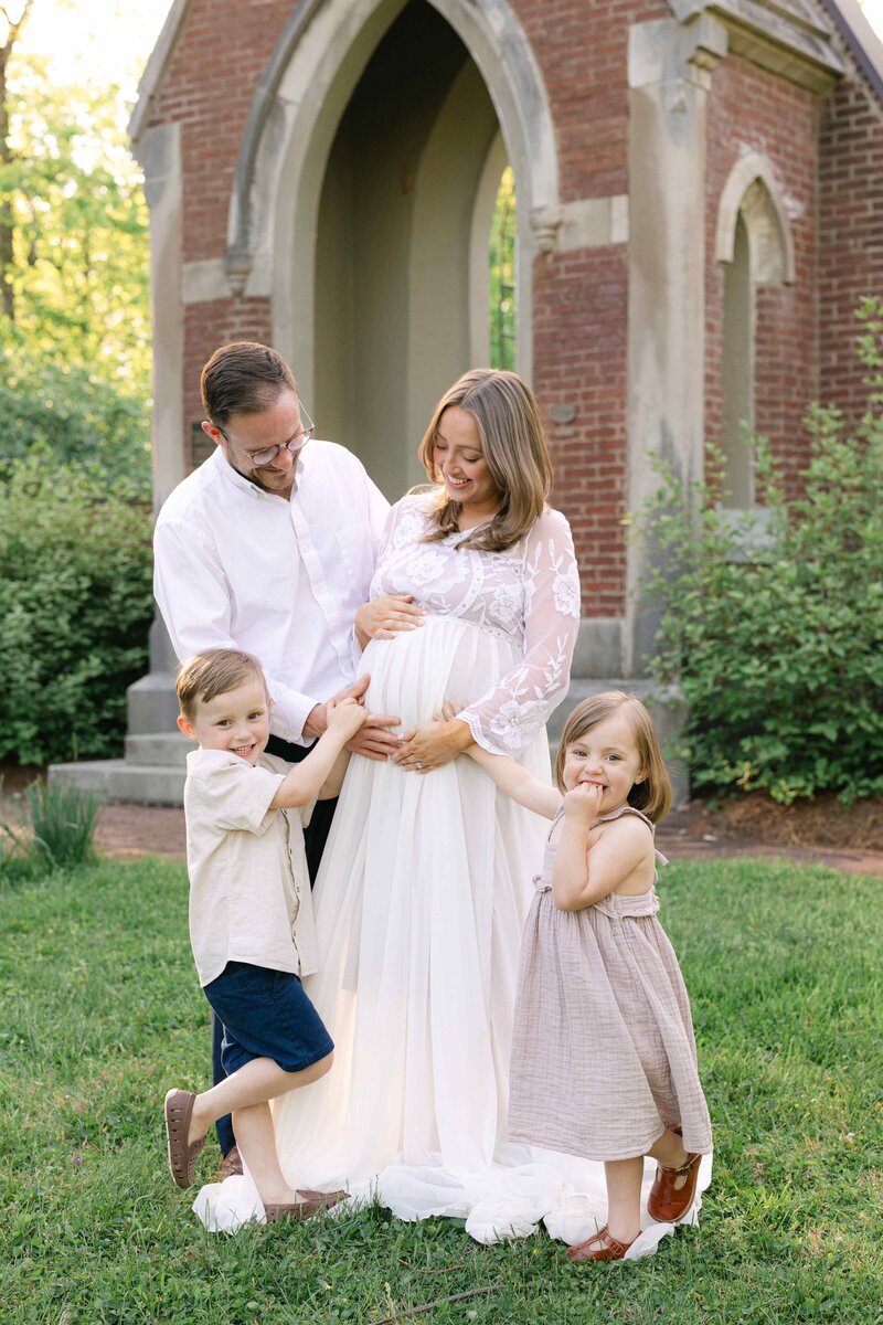 Portrait of a family of four all holding on to the pregnant mom's belly as they smile at each other in front of a brick structure taken by louisville maternity photographer missy marshall