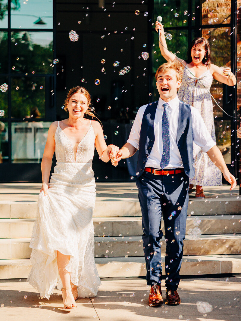 A joyful bride and groom holding hands and laughing as they walk outside amid floating bubbles, with a smiling woman in a floral dress behind them holding a microphone. Sunlight brightens the scene captured by a wedding photographer in Eau Claire.