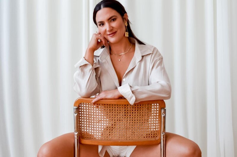 Emily Michele sitting in a beige oversized shirt, holding a woven bag — embodying calm confidence, grounded leadership, and effortless feminine strength.