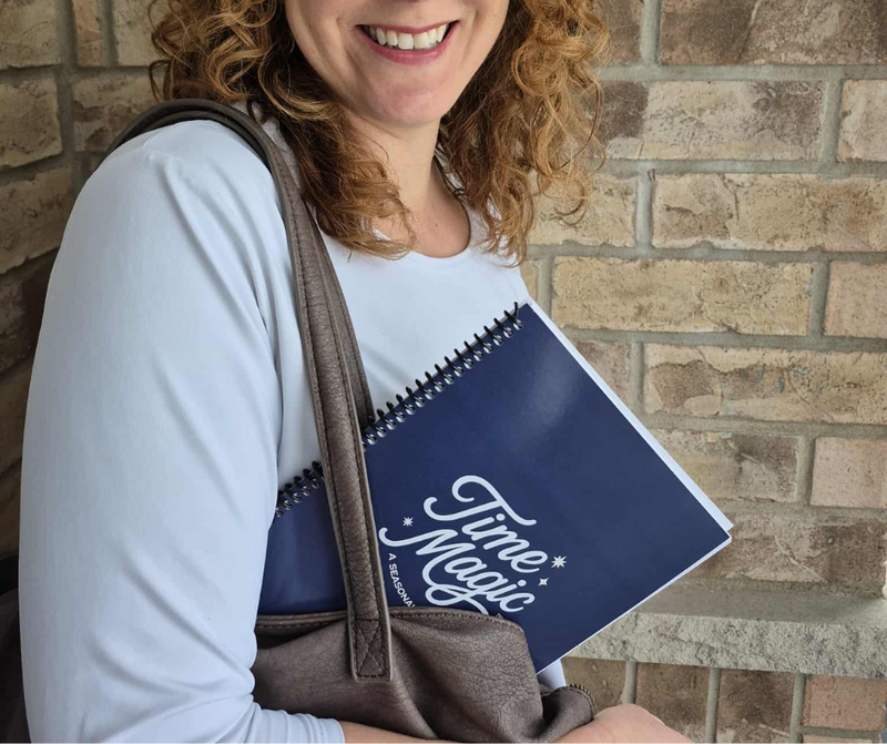 A woman smiling while holding the navy blue Time Magic Planner tucked under her arm, standing in front of a brick wall.