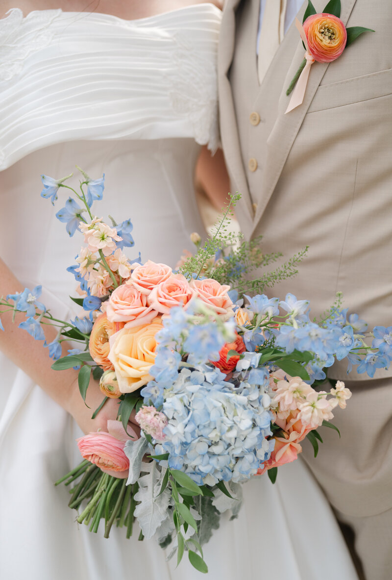 A bride holds a fresh flower bouquet of pinks, yellows, and blues with her groom in a tan suit.