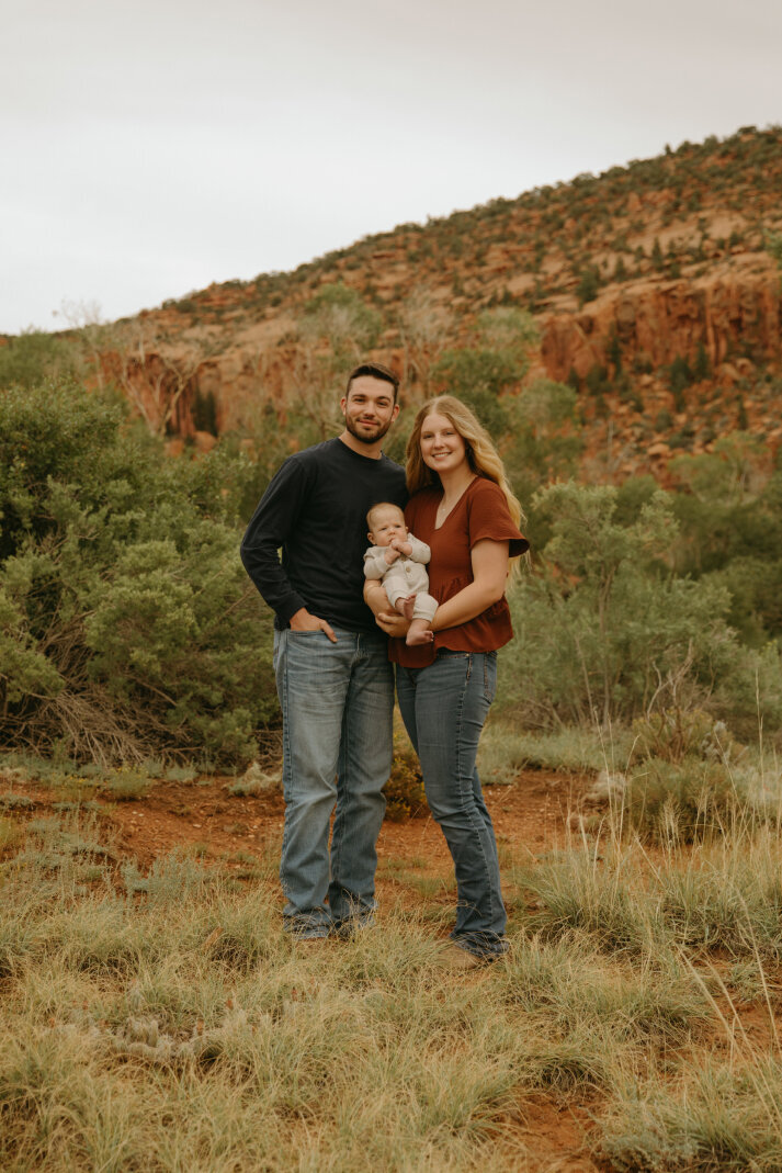 A family portrait of Hannah and her husband. She is holding her baby and they are in an outdoor setting in Southern Utah.