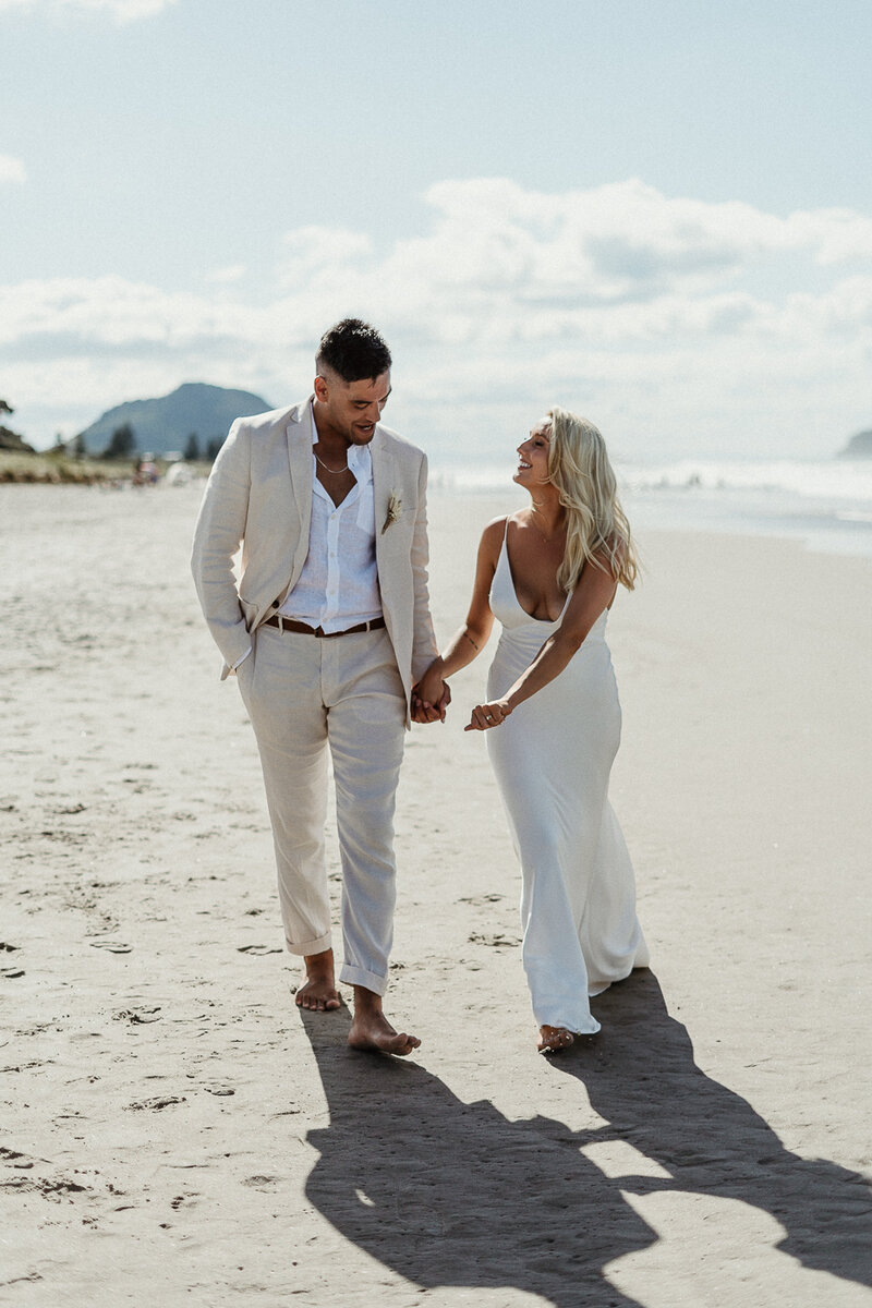 Just married and walking hand in hand along Mount Maunganui Beach in Tauranga, Bride wears beautiful trendy wedding dress and groom is in a cream suit