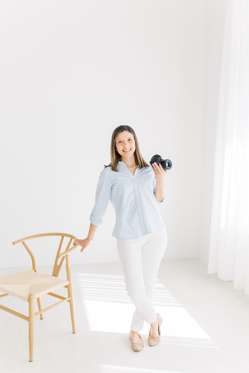 Austin, TX Family & Newborn Photography | A photographer leans against a chair in a white studio while holding her camera. 