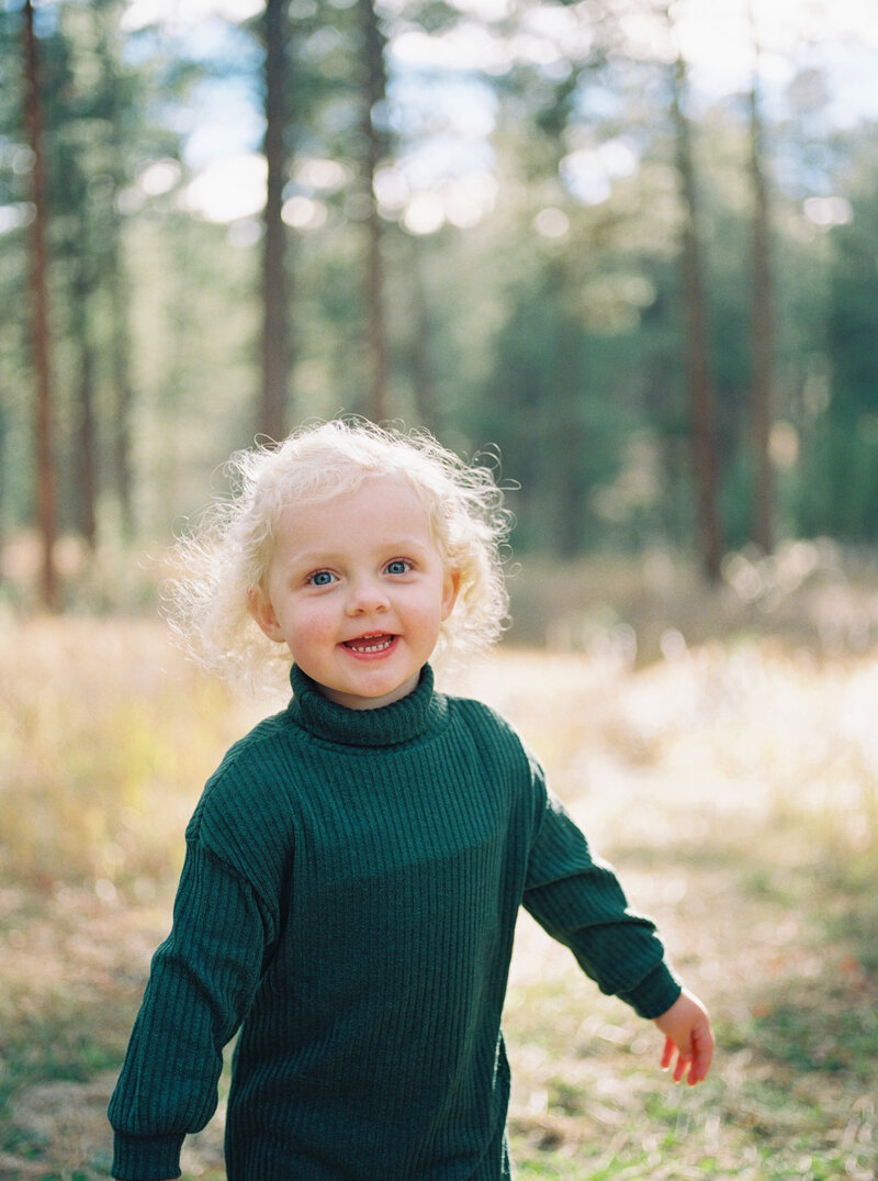 Blonde toddler with curly hair laughs at the camera while spinning in a field at sunset