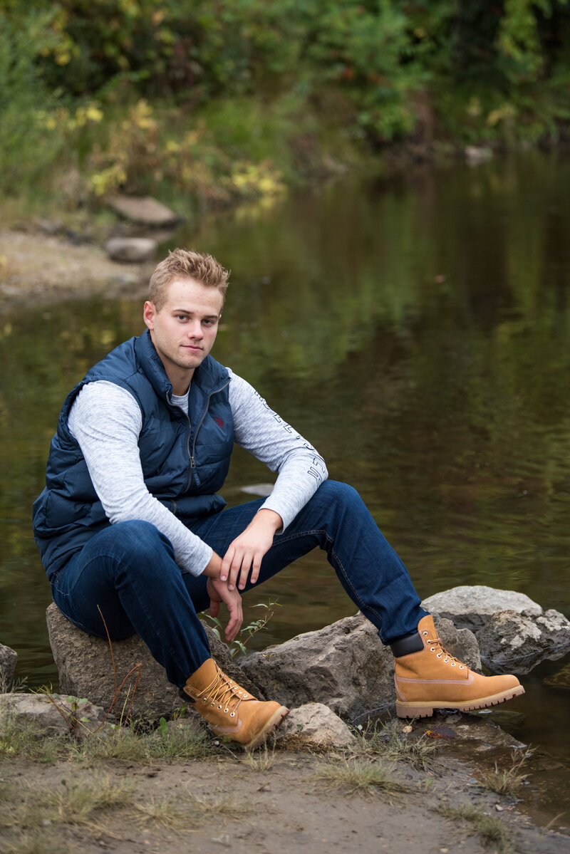 senior photo of guy in vest and jeans sitting on side of lake