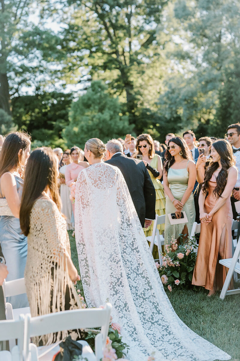 Rhode Island Wedding Photographers | A bride in a white lace gown walks down an outdoor aisle with guests in formal attire seated and standing, under sunny skies surrounded by greenery.