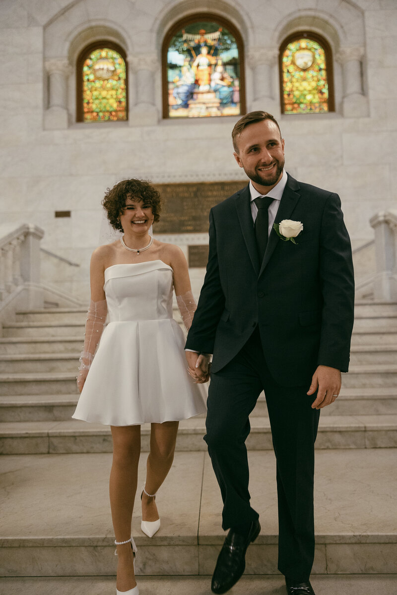 A bride in a short white dress and groom in a blue suit with white rose pinned to his chest smile while holding hands.