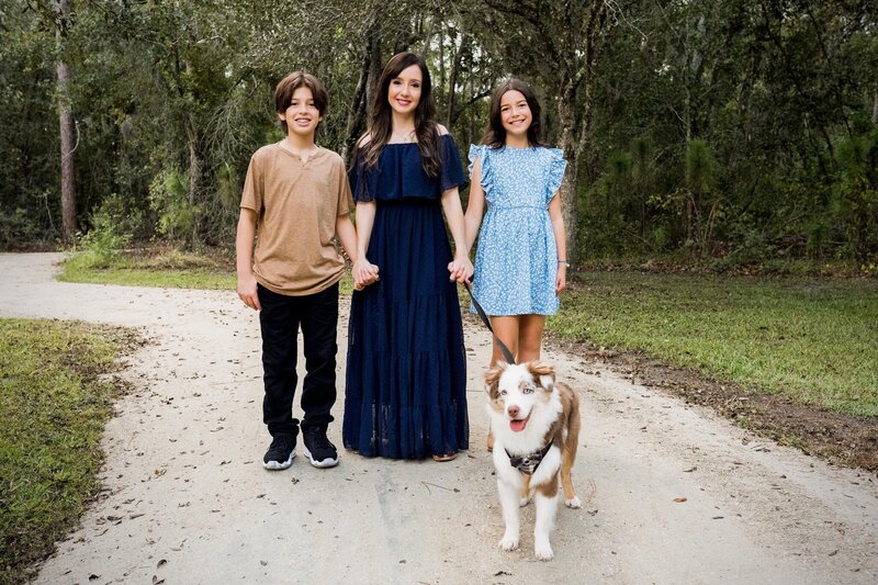A mother and her two kids play with their dog, surrounded by greenery and a clear blue sky.