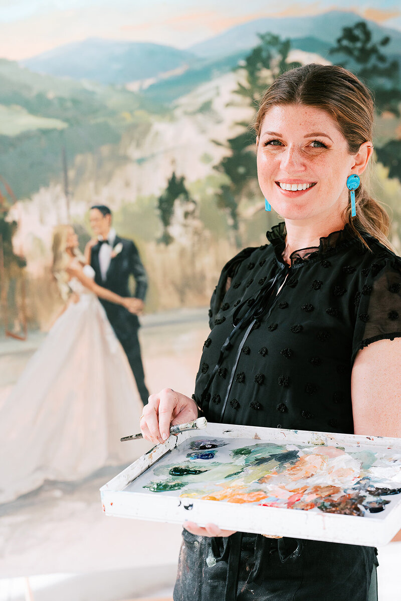 Stephanie wearing black shirt holding paint and brush with wedding painting of couple in background