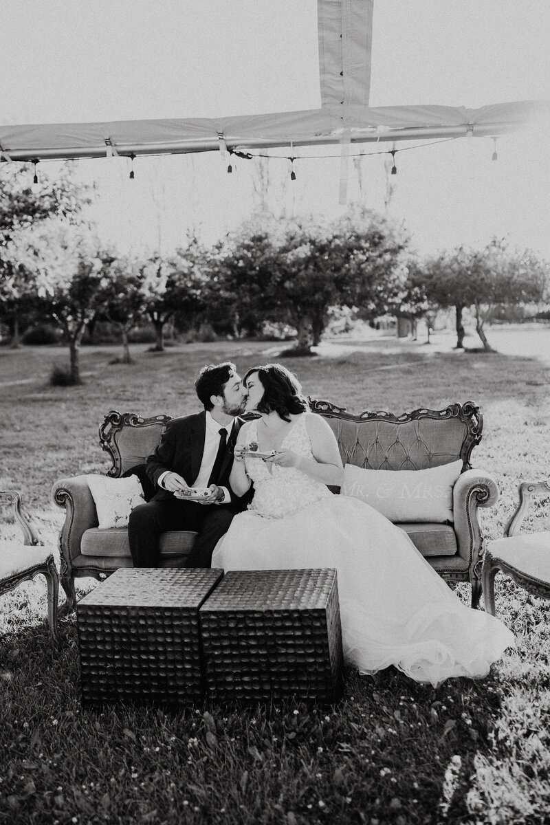 A bride and groom sit on a vintage couch outside in a cherry orchard