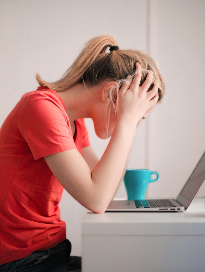 Woman with a red shirt and teal mug holding her head while looking frustrated at a laptop.