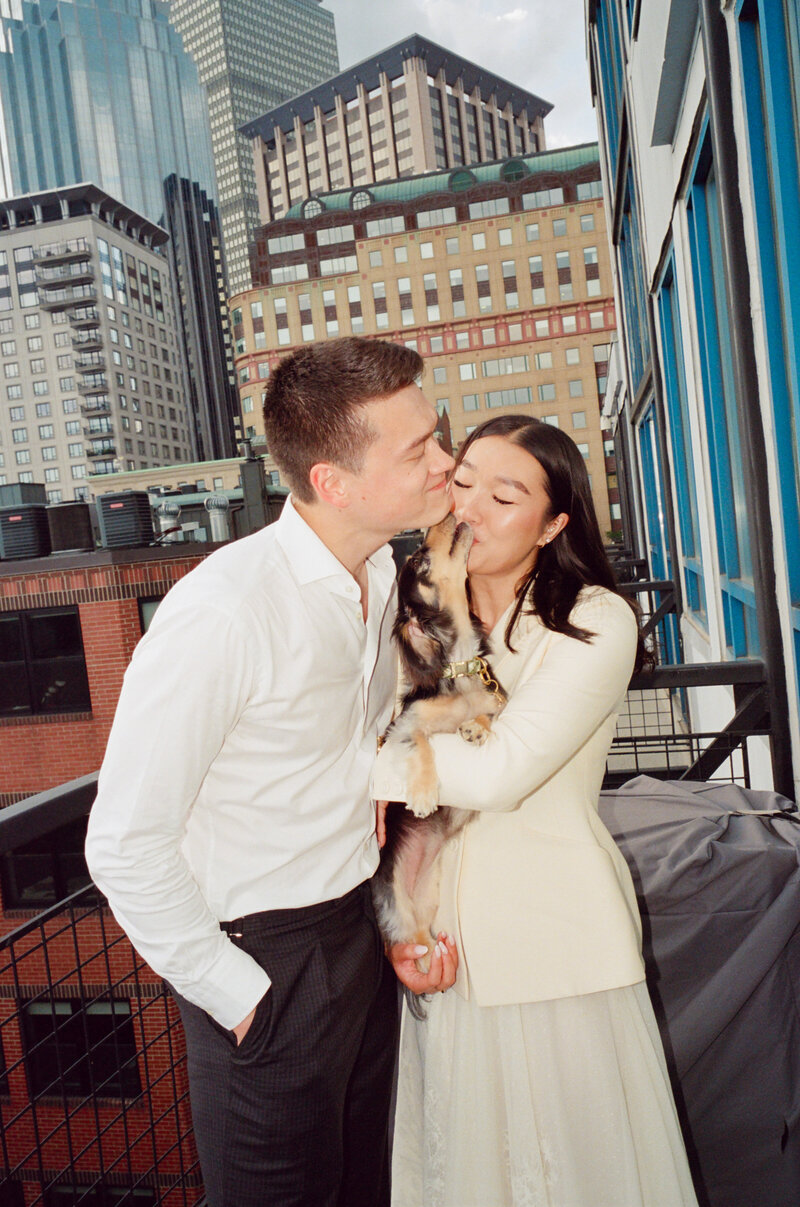 The dog, held in the bride and groom's arms, kisses them, backdropped by the Boston skyline. captured on film by a film wedding photographer at Castillo Holliday Photo + Film.