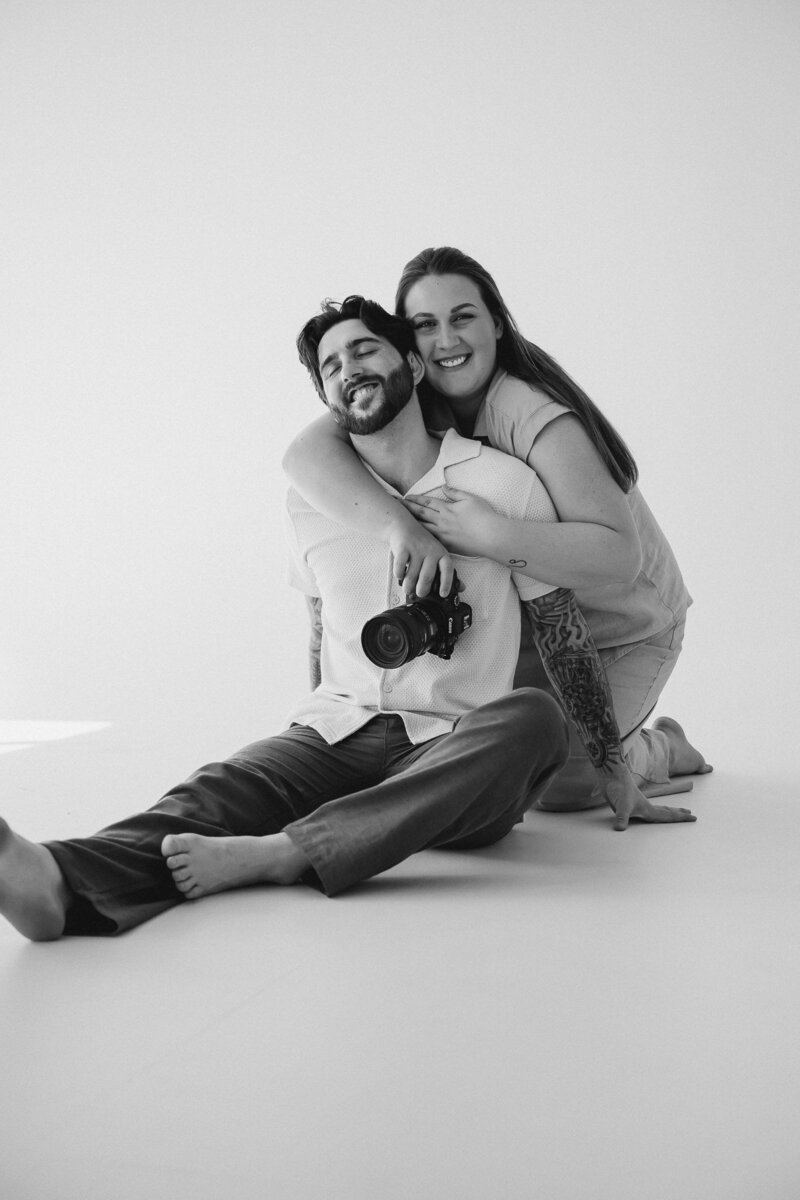 A person sitting on the ground with their partner leans over their shoulders and smiles