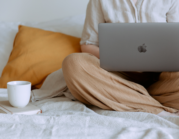 woman sitting cross-legged on a bed working on laptop with coffee next to her