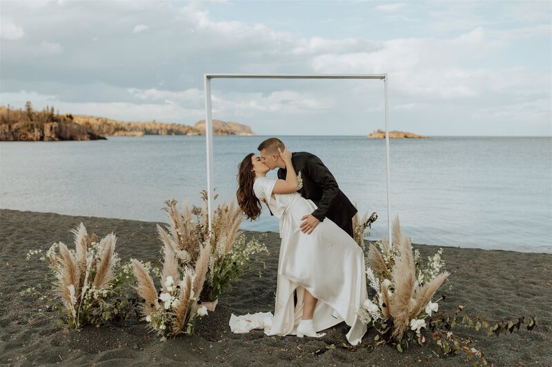 groom dipping bride and kissing under their ceremony arch on black beach 