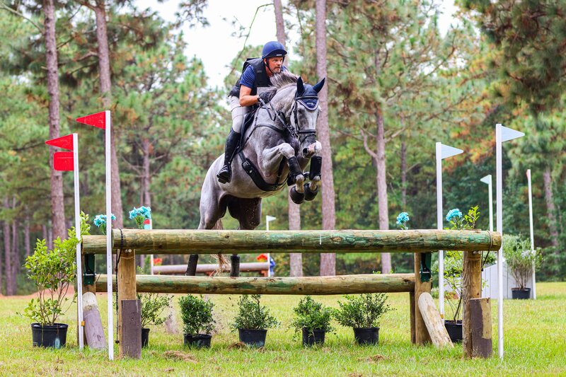 A grey horse jumping over a cross-country log fence during an eventing competition.
